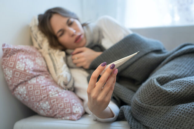Sick young Caucasian woman lying on sofa, wrapped in plaid blanket and scarf, having flu symptoms, holding and looking at thermometer, focus on thermometer/Cold, virus, sickness concept/Flu season